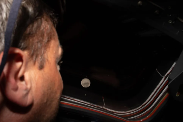 Artemis 2 commander Reid Wiseman gazes through the window of the Orion capsule Integrity at the Moon, appearing as a small bright dot against the darkness of deep space, during the historic lunar flyby on April 6, 2026.
