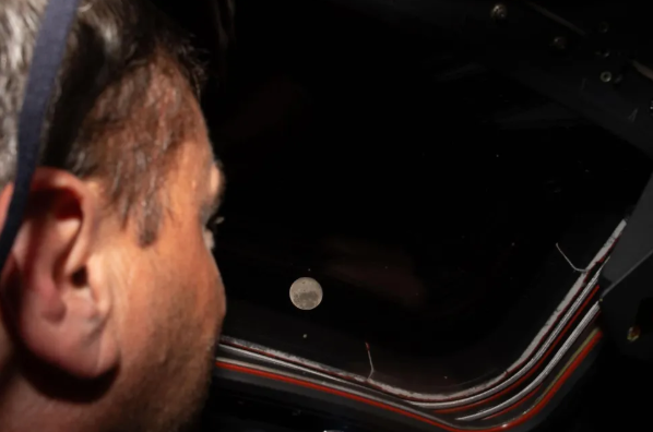 Artemis 2 commander Reid Wiseman gazes through the window of the Orion capsule Integrity at the Moon, appearing as a small bright dot against the darkness of deep space, during the historic lunar flyby on April 6, 2026.